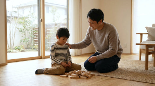崩れた積み木の前で落ち込む男の子の肩に手を置き、ひざをついて寄り添い、笑顔で声をかける大人の男性