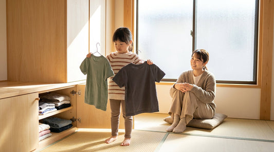 2枚のTシャツを手に持って、どちらを着るか選ぶ女の子と、少し離れて座って見守る大人の女性
