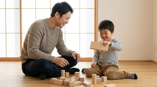 リビングの床で、大人の男性と男の子が木の積み木を手に取って笑顔で遊んでいる様子