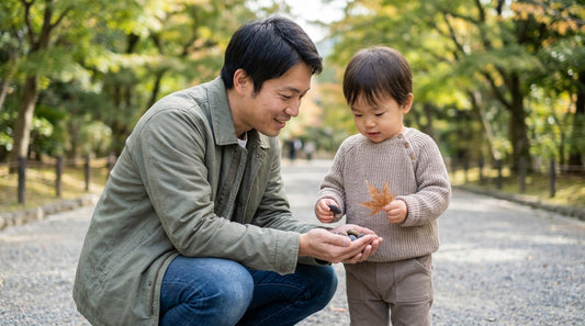 公園の小道でしゃがみ、手のひらにのせた小石を子どもと一緒に見る大人の男性と、落ち葉を持つ2歳くらいの子ども